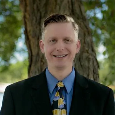 Chad Eisinger — professional headshot in navy suit and blue shirt, smiling in a bright office