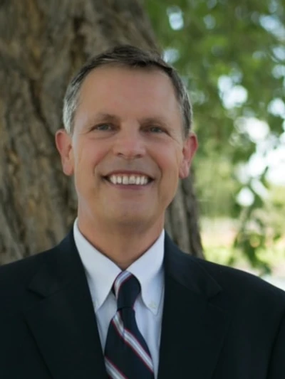 Tim D. Binegar smiling outdoors in a navy suit, white shirt, and striped tie